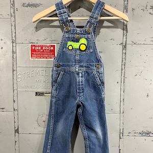 May include: Blue denim overalls with a green and yellow tractor patch on the front. The overalls have adjustable straps and a button closure.