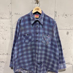Pu&ograve; includere: Una camicia a maniche lunghe con bottoni e un motivo a quadri blu e azzurro. La camicia presenta un colletto a punta, due tasche sul petto con chiusura a bottone e una serie di bottoni neri sul davanti. Sono presenti segni di usura visibili.