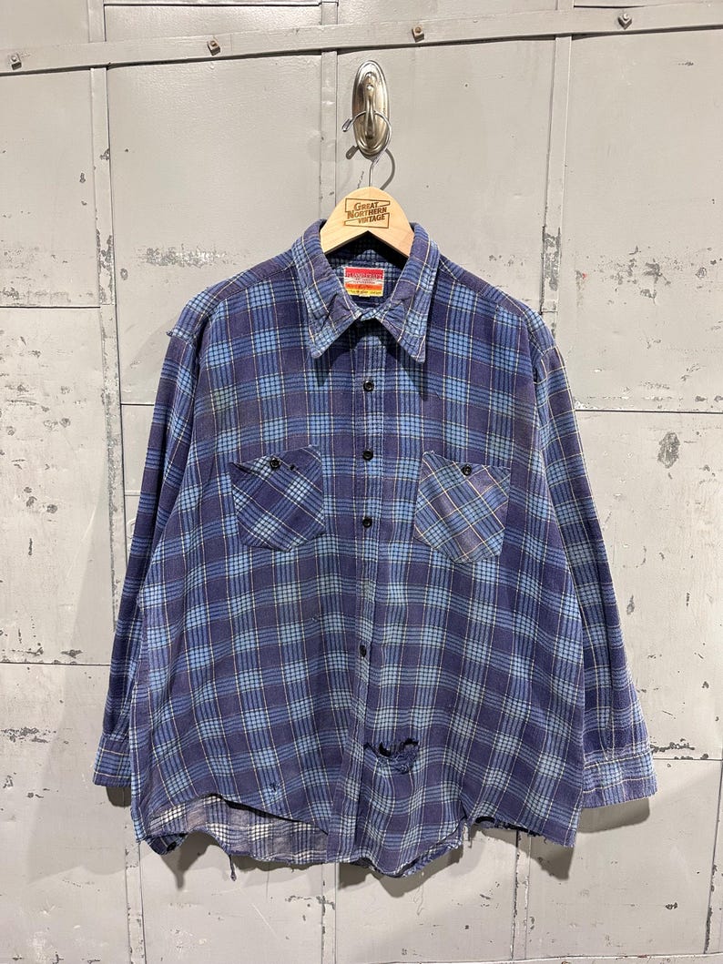 Pu&ograve; includere: Una camicia a maniche lunghe con bottoni e un motivo a quadri blu e azzurro. La camicia presenta un colletto a punta, due tasche sul petto con chiusura a bottone e una serie di bottoni neri sul davanti. Sono presenti segni di usura visibili.