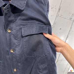 May include: Navy blue jacket with a collared neckline, button-up front, and two front pockets. The jacket has a classic design and is made of a durable fabric. The brand label is visible at the top.
