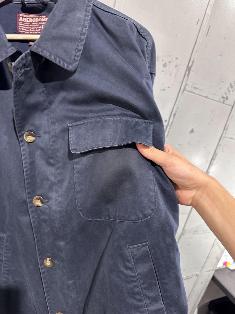 May include: Navy blue jacket with a collared neckline, button-up front, and two front pockets. The jacket has a classic design and is made of a durable fabric. The brand label is visible at the top.