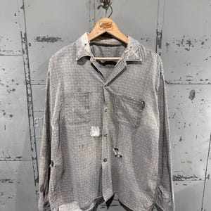 May include: A worn grey button-down shirt with a small geometric pattern, two chest pockets, and signs of wear, including holes and patches. The shirt hangs on a wooden hanger with the text "Great Northern Vintage".