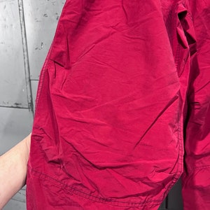 May include: A pair of red trousers made from a lightweight, crinkled fabric. The trousers have a simple design with a single seam down the front and back of each leg.