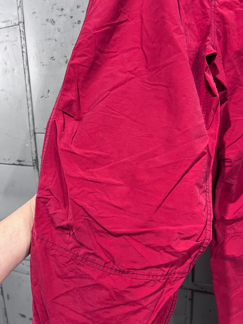 May include: A pair of red trousers made from a lightweight, crinkled fabric. The trousers have a simple design with a single seam down the front and back of each leg.