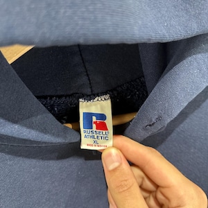 May include: Close-up of a navy blue hoodie with a hood. The tag reads "RUSSELL ATHLETIC XL MADE IN MEXICO" with a red and blue logo. The hoodie is made of a soft, comfortable material.