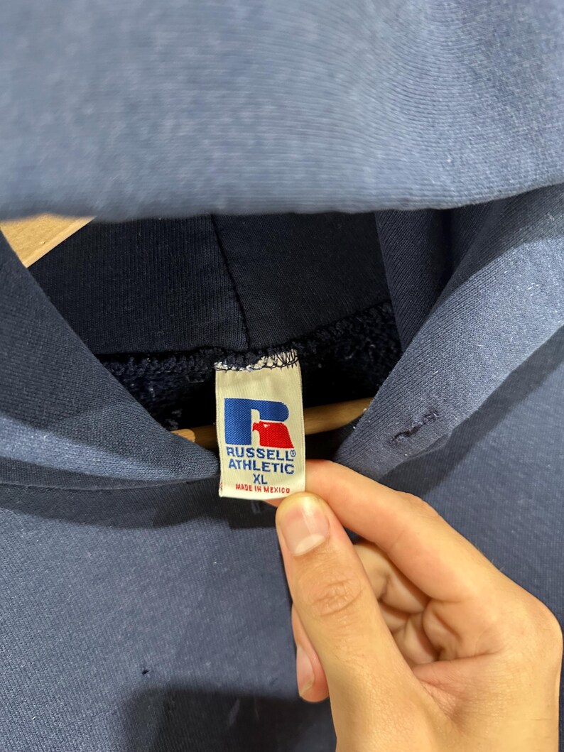May include: Close-up of a navy blue hoodie with a hood. The tag reads "RUSSELL ATHLETIC XL MADE IN MEXICO" with a red and blue logo. The hoodie is made of a soft, comfortable material.