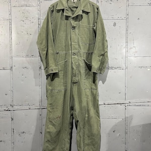 May include: A green utility jumpsuit with a button-up front and multiple pockets. The jumpsuit is hanging on a hanger against a white wall.