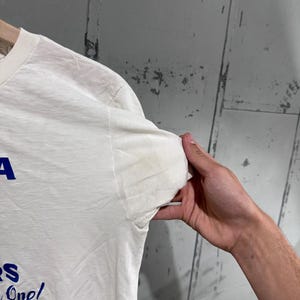 May include: A white t-shirt with blue text that reads "A" and "S One!" The shirt is held up by a person's hand. The shirt has a stain on the sleeve.
