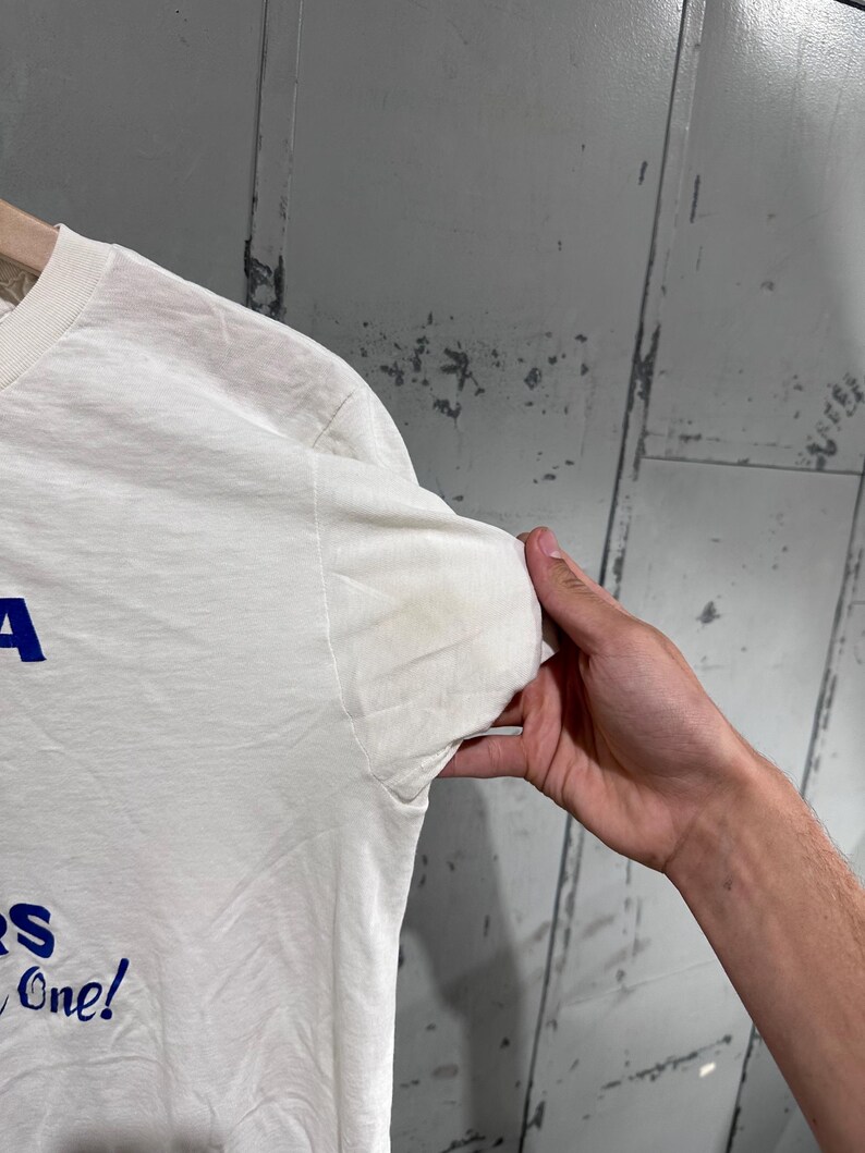 May include: A white t-shirt with blue text that reads "A" and "S One!" The shirt is held up by a person's hand. The shirt has a stain on the sleeve.