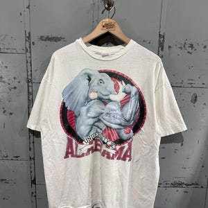 May include: A white short-sleeve t-shirt featuring a graphic of a muscular grey elephant flexing its arm with the text "NO MERCY" on its bicep. Below the elephant, a banner reads "UNIVERSITY OF ALABAMA" in red lettering over a circular red background.