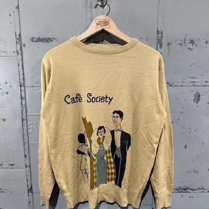 May include: Tan-colored sweater with the words "Cafe Society" and a graphic of three people in formal attire. The sweater is hanging on a wooden hanger against a gray metal background.