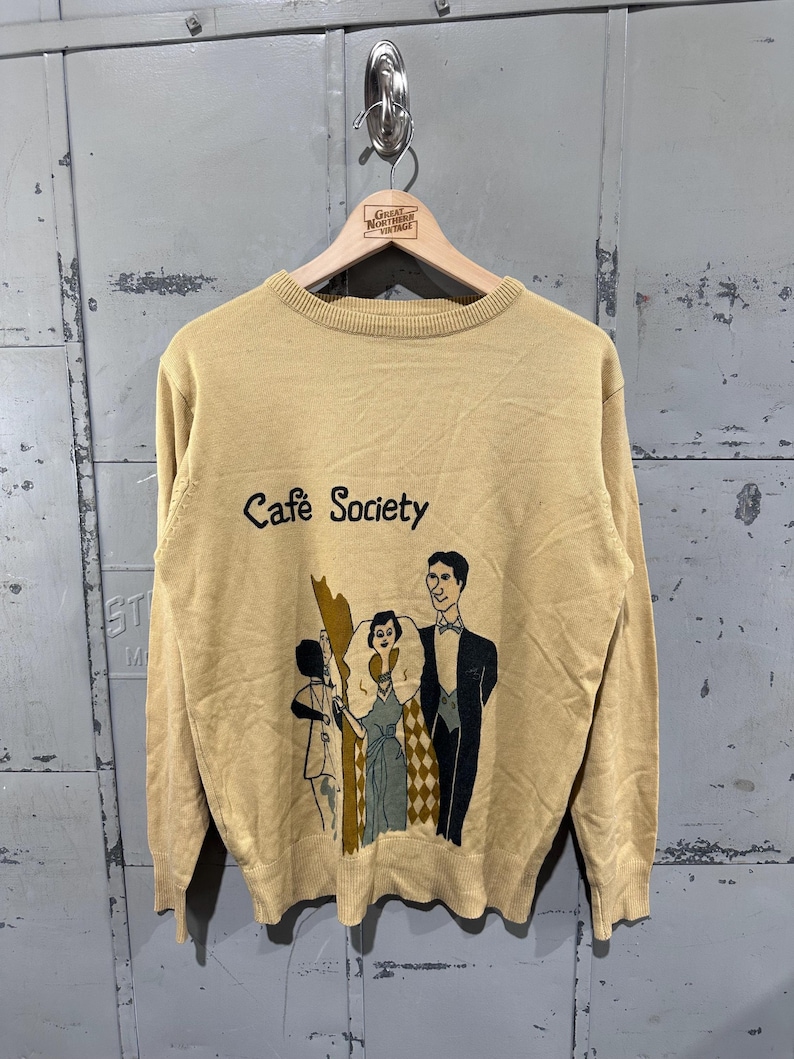 May include: Tan-colored sweater with the words "Cafe Society" and a graphic of three people in formal attire. The sweater is hanging on a wooden hanger against a gray metal background.