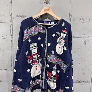 May include: Navy blue cardigan sweater with embroidered snowmen and snowflakes. The sweater features a button-up front and a decorative border with red berries and green leaves. A cozy winter garment.