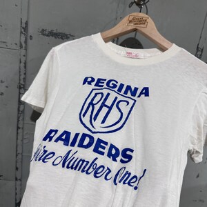 May include: White t-shirt with blue text and graphics. The text reads "REGINA RHS RAIDERS We're Number One!" The shirt is hanging on a wooden hanger with a "Great Northern Vintage" label.