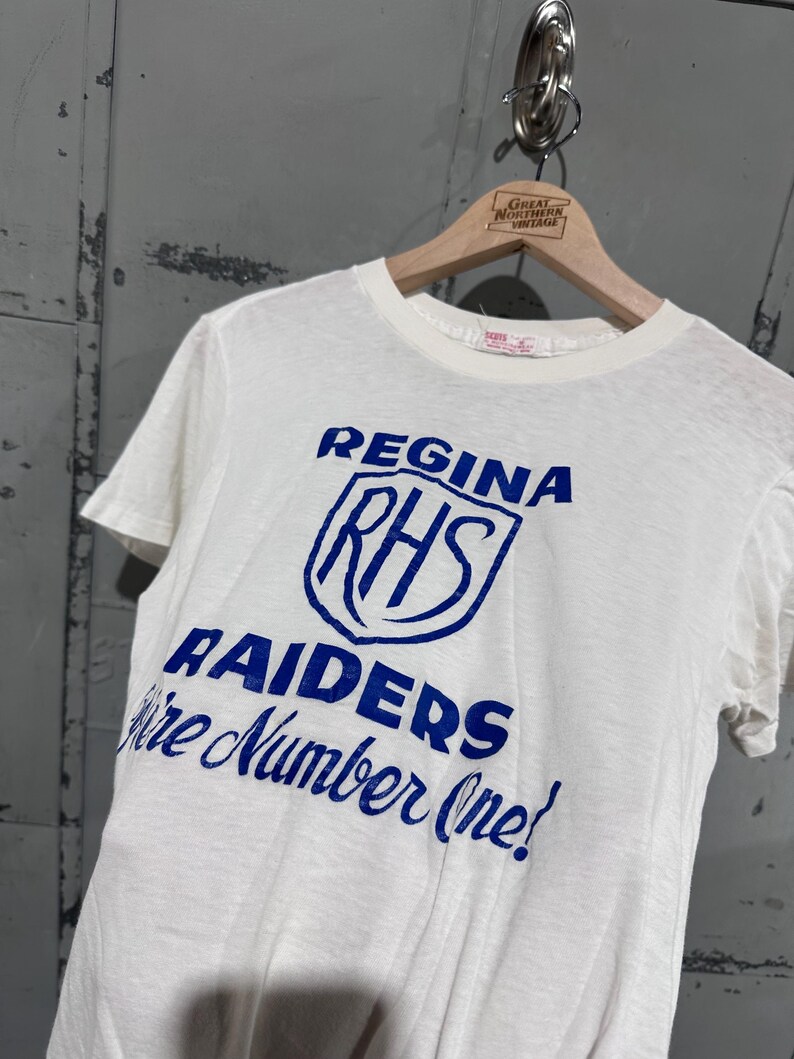 May include: White t-shirt with blue text and graphics. The text reads "REGINA RHS RAIDERS We're Number One!" The shirt is hanging on a wooden hanger with a "Great Northern Vintage" label.