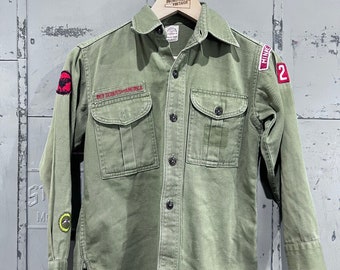May include: A green Boy Scout shirt with long sleeves and a button-up front. The shirt has two pockets on the front and a patch that says "Boy Scouts of America". There are also patches on the sleeves, one with a red eagle and the other with the text "Troop 2".