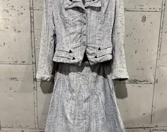 Puede incluir: Un traje vintage gris claro con chaqueta y falda. La chaqueta tiene cuello con solapas, cierre de botones y detalles decorativos negros. La falda es de largo midi. El traje está colgado en una percha de madera.