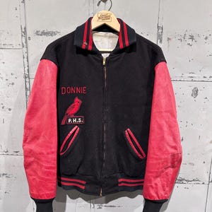 Op de afbeelding: Zwarte vintage varsity jas met rode mouwen. De jas heeft de naam "DONNIE" en een kardinaalvogel patch. De letters "P.H.S." staan ook op de jas. Het heeft een zwarte kraag met rode strepen en een ritssluiting.