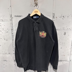 May include: Black long-sleeved polo shirt with a collared neckline and three-button placket. Features an embroidered crest on the left chest with gold, red, and yellow accents. The shirt is hanging on a wooden hanger.