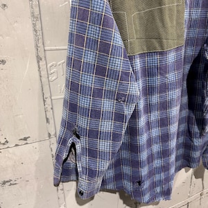 Pu&ograve; includere: Una camicia a maniche lunghe con bottoni caratterizzata da un motivo a quadri blu e viola con sottili linee gialle. La camicia presenta una toppa rettangolare verde oliva sul petto e segni di usura con piccoli fori sfilacciati sulla manica.