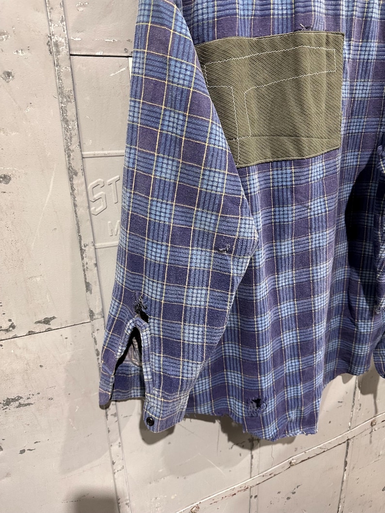 Pu&ograve; includere: Una camicia a maniche lunghe con bottoni caratterizzata da un motivo a quadri blu e viola con sottili linee gialle. La camicia presenta una toppa rettangolare verde oliva sul petto e segni di usura con piccoli fori sfilacciati sulla manica.