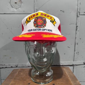 May include: A red and white trucker hat with the text "FIRE DEPT." and "YOUR CUSTOM COPY HERE." The hat features a red brim and a yellow emblem. The hat is displayed on a glass head.