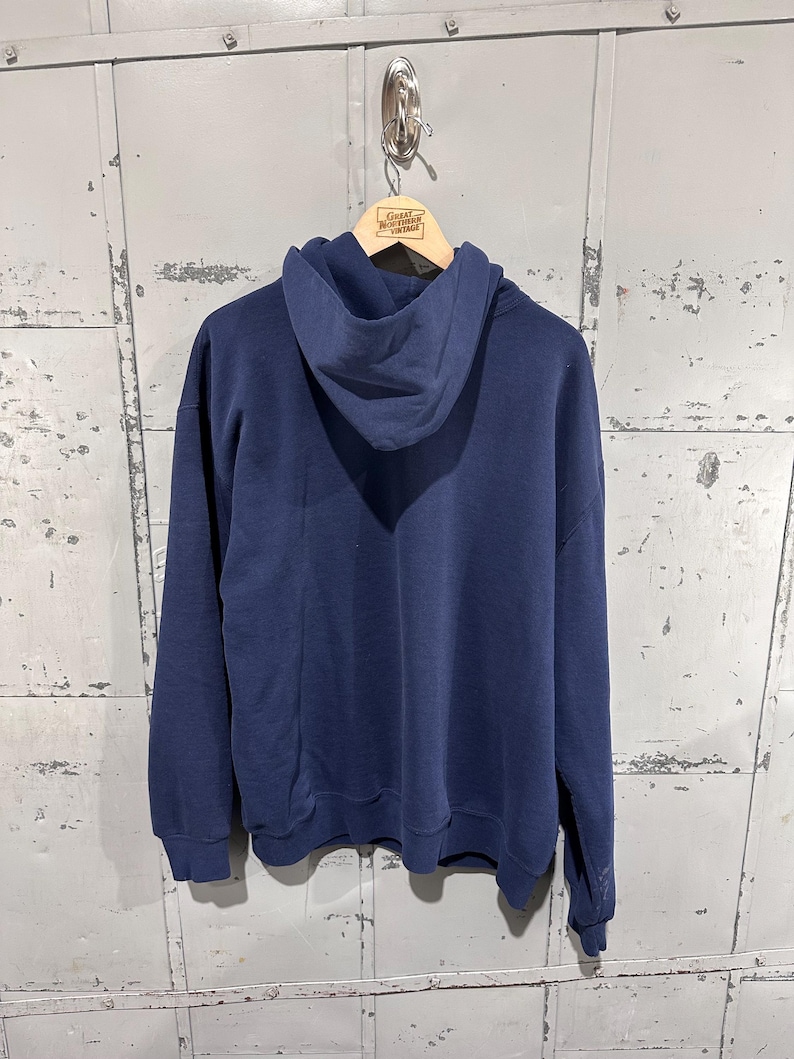 May include: A navy blue hoodie with a drawstring hood and long sleeves. The hoodie is hanging on a wooden hanger. The tag on the hanger reads "Great Northern Vintage".
