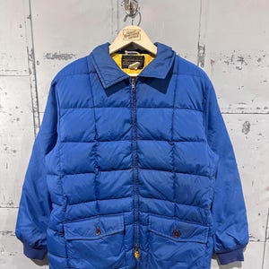 Pode incluir: Casaco acolchoado azul vintage com interior amarelo e fecho de correr. O casaco tem dois bolsos frontais com botões e uma gola. A etiqueta diz "Great Northern Vintage".