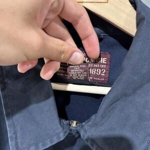 May include: A navy blue jacket with a zipper closure. The jacket has a label that reads "Abercrombie 1892" and includes washing instructions. The jacket is hanging on a wooden hanger with the text "Great Northern Vintage".