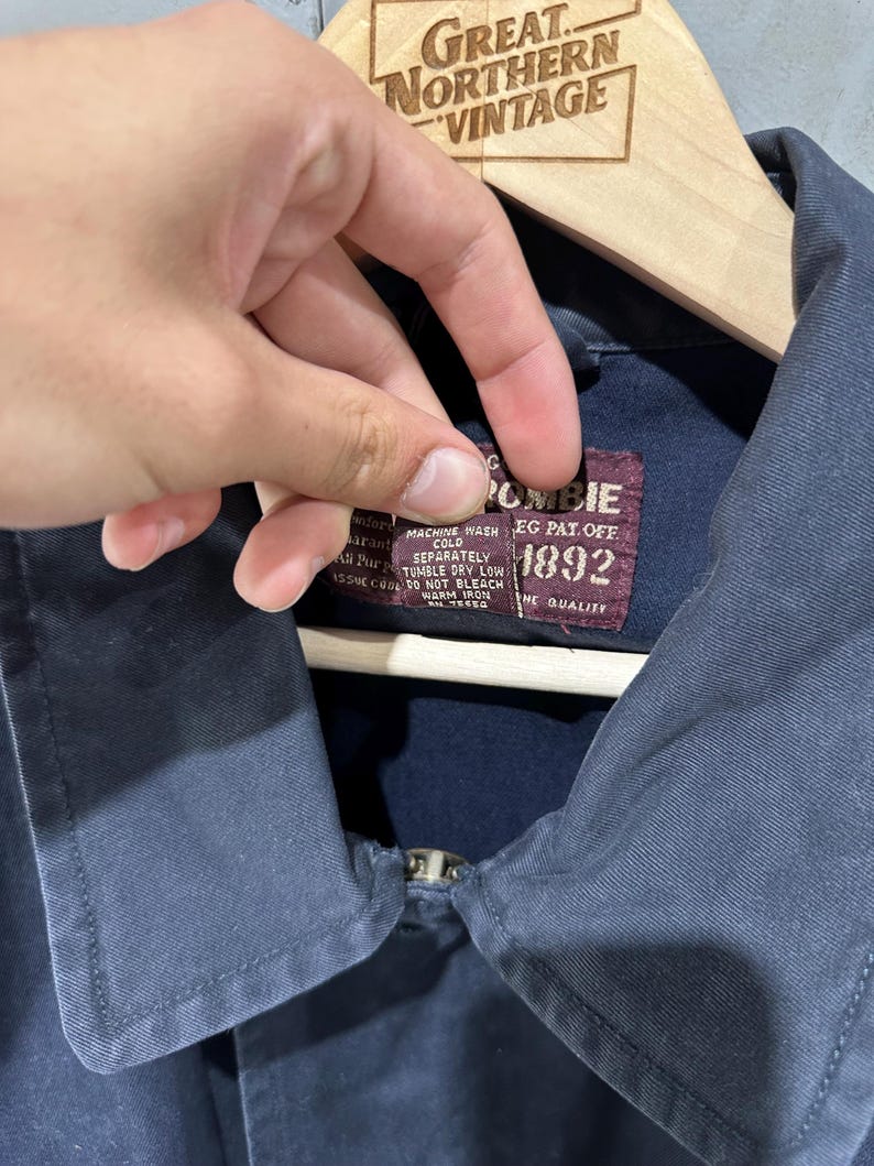 May include: A navy blue jacket with a zipper closure. The jacket has a label that reads "Abercrombie 1892" and includes washing instructions. The jacket is hanging on a wooden hanger with the text "Great Northern Vintage".