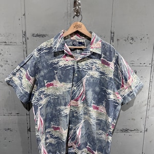 Pode incluir: Camisa de manga curta com botões e estampa náutica com veleiros em um fundo azul e cinza. A camisa está pendurada em um cabide de madeira. A camisa tem uma gola clássica e um ajuste relaxado.