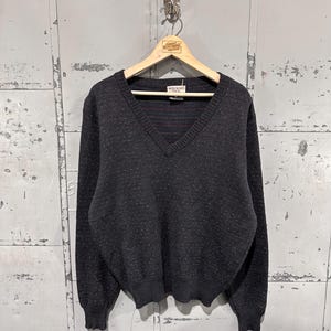 May include: A dark gray V-neck sweater with a speckled pattern. The sweater is hanging on a wooden hanger. The tag reads "Wildebond USA". The background is a gray metal wall.