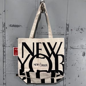 May include: A cream-colored canvas tote bag featuring large, bold black lettering that spells out NEW YORKER. The design includes the smaller text THE NEW YORKER centered within the letter O. The bag has two long fabric handles for carrying.
