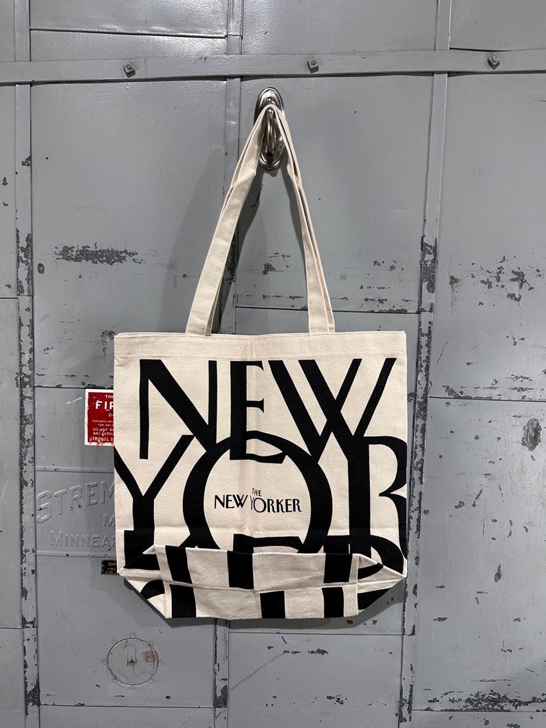 May include: A cream-colored canvas tote bag featuring large, bold black lettering that spells out NEW YORKER. The design includes the smaller text THE NEW YORKER centered within the letter O. The bag has two long fabric handles for carrying.