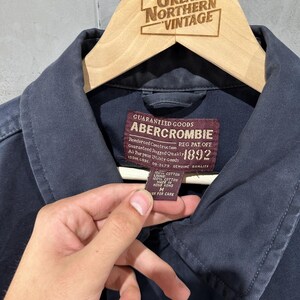 May include: Close-up of a navy blue Abercrombie jacket. The jacket is hanging on a wooden hanger with the text "Great Northern Vintage" at the top. The jacket has a label with text including "Abercrombie" and "1892".