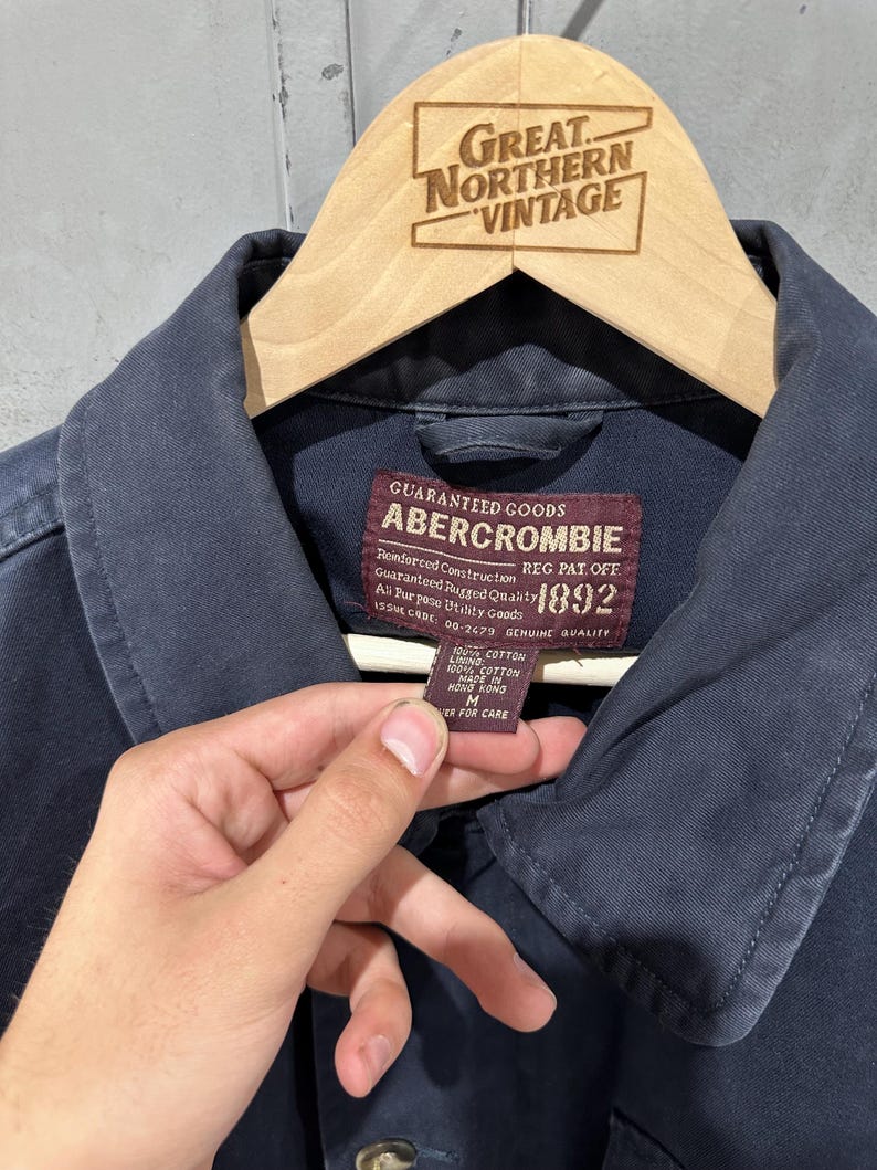 May include: Close-up of a navy blue Abercrombie jacket. The jacket is hanging on a wooden hanger with the text "Great Northern Vintage" at the top. The jacket has a label with text including "Abercrombie" and "1892".