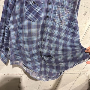 Pu&ograve; includere: Una camicia a maniche lunghe con bottoni caratterizzata da un motivo a quadri blu e blu navy con sottili linee gialle. La camicia presenta due tasche sul petto, una chiusura frontale con bottoni e segni di usura, tra cui un foro e un orlo sfilacciato.