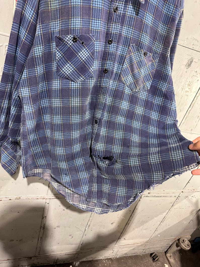 Pu&ograve; includere: Una camicia a maniche lunghe con bottoni caratterizzata da un motivo a quadri blu e blu navy con sottili linee gialle. La camicia presenta due tasche sul petto, una chiusura frontale con bottoni e segni di usura, tra cui un foro e un orlo sfilacciato.