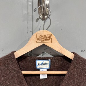 K&ouml;nnte beinhalten: Ein brauner V-Ausschnitt-Pullover mit texturierter Strickoberfl&auml;che. Das Kleidungsst&uuml;ck hat ein blau-wei&szlig;es Pendleton Originals-Etikett am inneren Halsausschnitt sowie ein zweites Etikett mit der Gr&ouml;&szlig;e XL und 100% Wolle, h&auml;ngend an einem Holzkleiderb&uuml;gel.