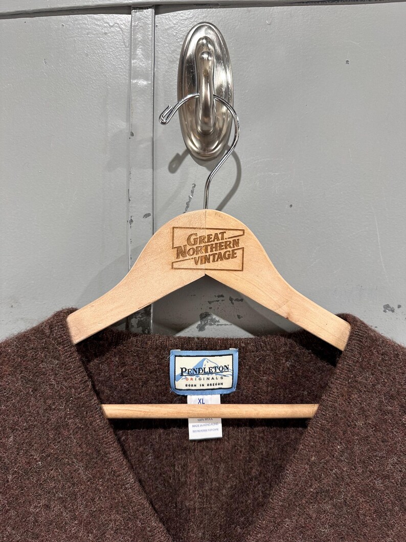 K&ouml;nnte beinhalten: Ein brauner V-Ausschnitt-Pullover mit texturierter Strickoberfl&auml;che. Das Kleidungsst&uuml;ck hat ein blau-wei&szlig;es Pendleton Originals-Etikett am inneren Halsausschnitt sowie ein zweites Etikett mit der Gr&ouml;&szlig;e XL und 100% Wolle, h&auml;ngend an einem Holzkleiderb&uuml;gel.