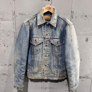 Könnte beinhalten: Eine verwaschene blaue Jeansjacke mit spitzem Kragen, langen Ärmeln mit geknöpften Manschetten und einer Knopfleiste vorne. Die Jacke verfügt über zwei Brusttaschen mit geknöpften Klappen, ein kleines rotes Etikett und eine abgenutzte Optik.