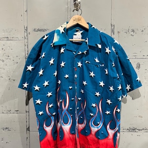 May include: A short-sleeved button-down shirt with a vibrant design. The shirt features a dark blue background adorned with white stars and a flame pattern in red, white, and blue along the bottom. The collar has a faded blue pattern.