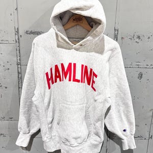 Może przedstawiać: Jasnoszara bluza z kapturem z napisem "HAMLINE" w czerwonych literach na piersi. Bluza posiada przednią kieszeń, kaptur ze sznurkiem i prążkowane mankiety. Małe logo Champion jest widoczne na lewym rękawie.