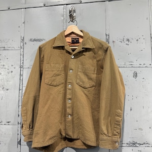 May include: A tan button-down shirt with two chest pockets and long sleeves. The shirt is hanging on a wooden hanger against a metallic gray background. The shirt has a classic collar and buttoned cuffs.