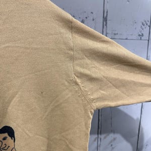 May include: Tan long-sleeved sweater featuring a cartoon illustration of a man in a tuxedo. The sweater has a ribbed texture and a crew neck. The illustration is in black and white.