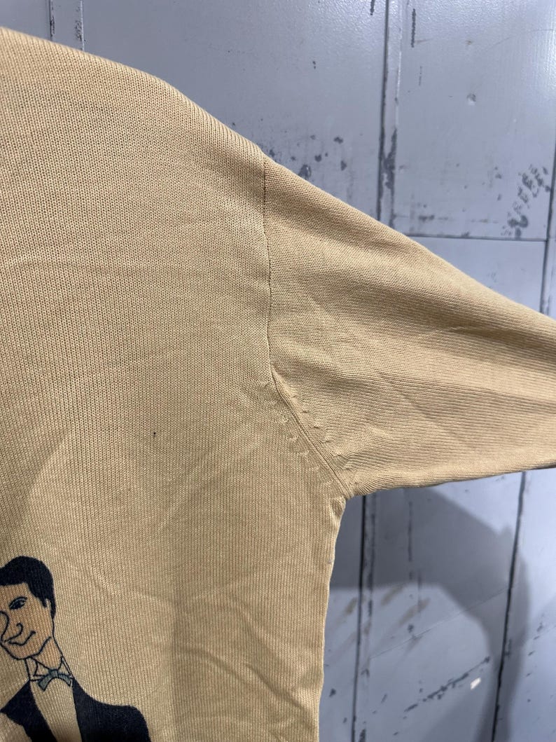 May include: Tan long-sleeved sweater featuring a cartoon illustration of a man in a tuxedo. The sweater has a ribbed texture and a crew neck. The illustration is in black and white.