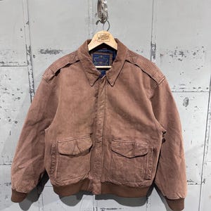 May include: A brown suede bomber jacket with two front pockets and a ribbed hem and cuffs. The jacket has a collared neckline and shoulder epaulets. The jacket is hanging on a wooden hanger.