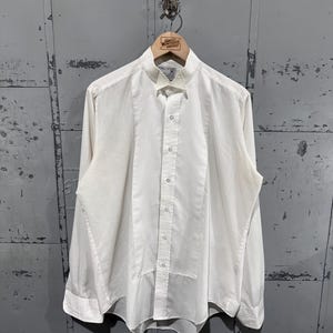 Pode incluir: Uma camisa de smoking branca de manga comprida com um painel frontal plissado, gola de bico e carcela central com botões. A camisa é feita de tecido liso e inclui punhos clássicos, apresentando um design formal.