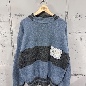 May include: A blue and gray sweater with a crew neck and a horizontal gray stripe across the chest. The sweater has a small patch with text and graphics. The sweater is hanging on a wooden hanger.