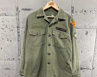 May include: Olive green military shirt with two chest pockets and button closures. The shirt features "U.S. ARMY" text above the left pocket and a patch on the right sleeve. The shirt is hanging on a wooden hanger.